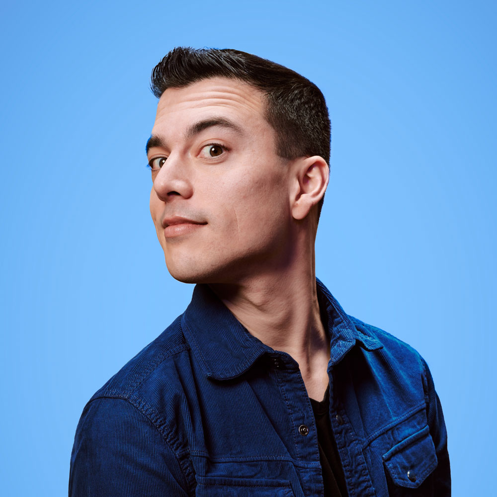 Cabaretier Andries Tunru kijkt eigenzinnig schuin omhoog voor een blauwe achtergrond.