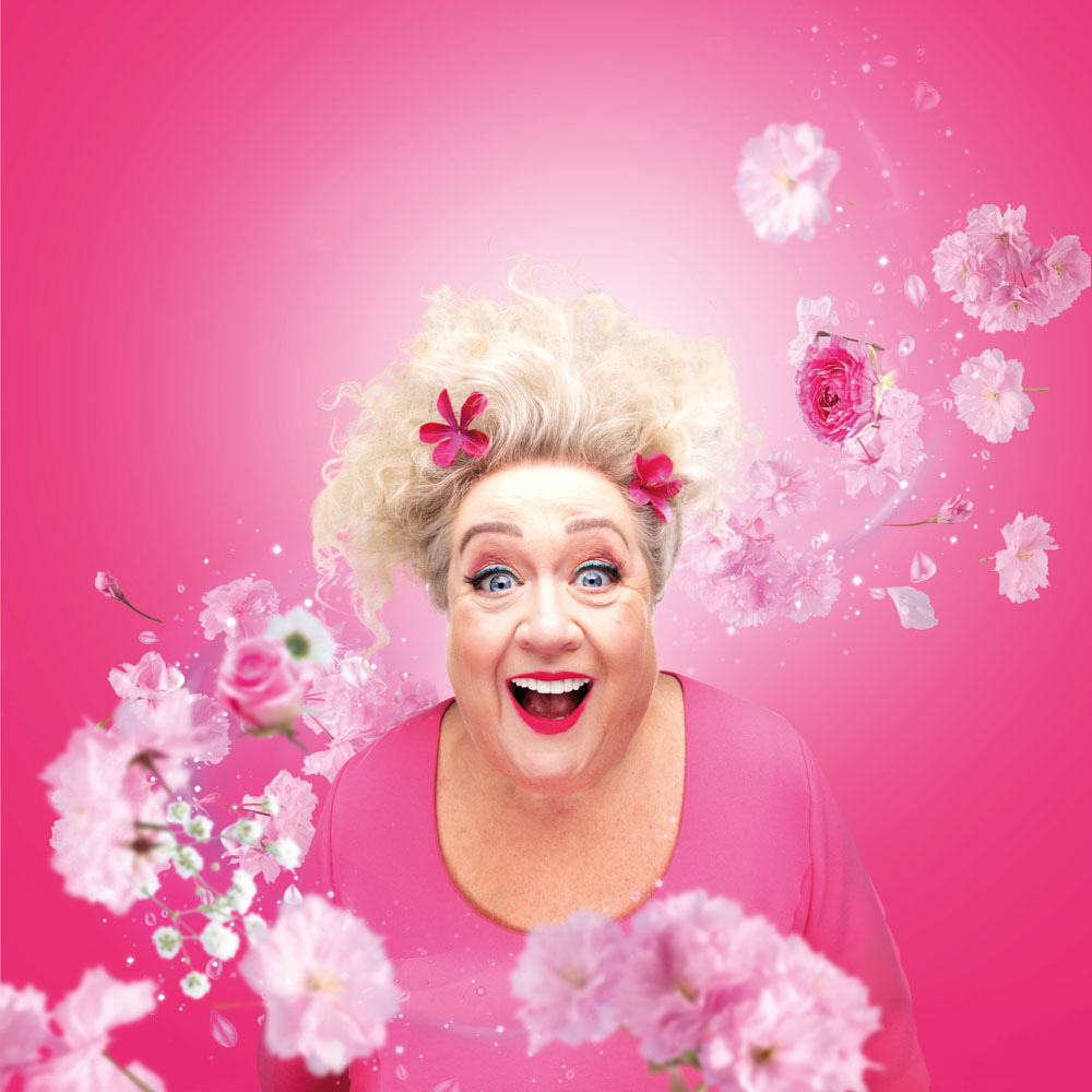 Christel de Laat lacht uitbundig voor een felroze achtergrond vol bloemen.