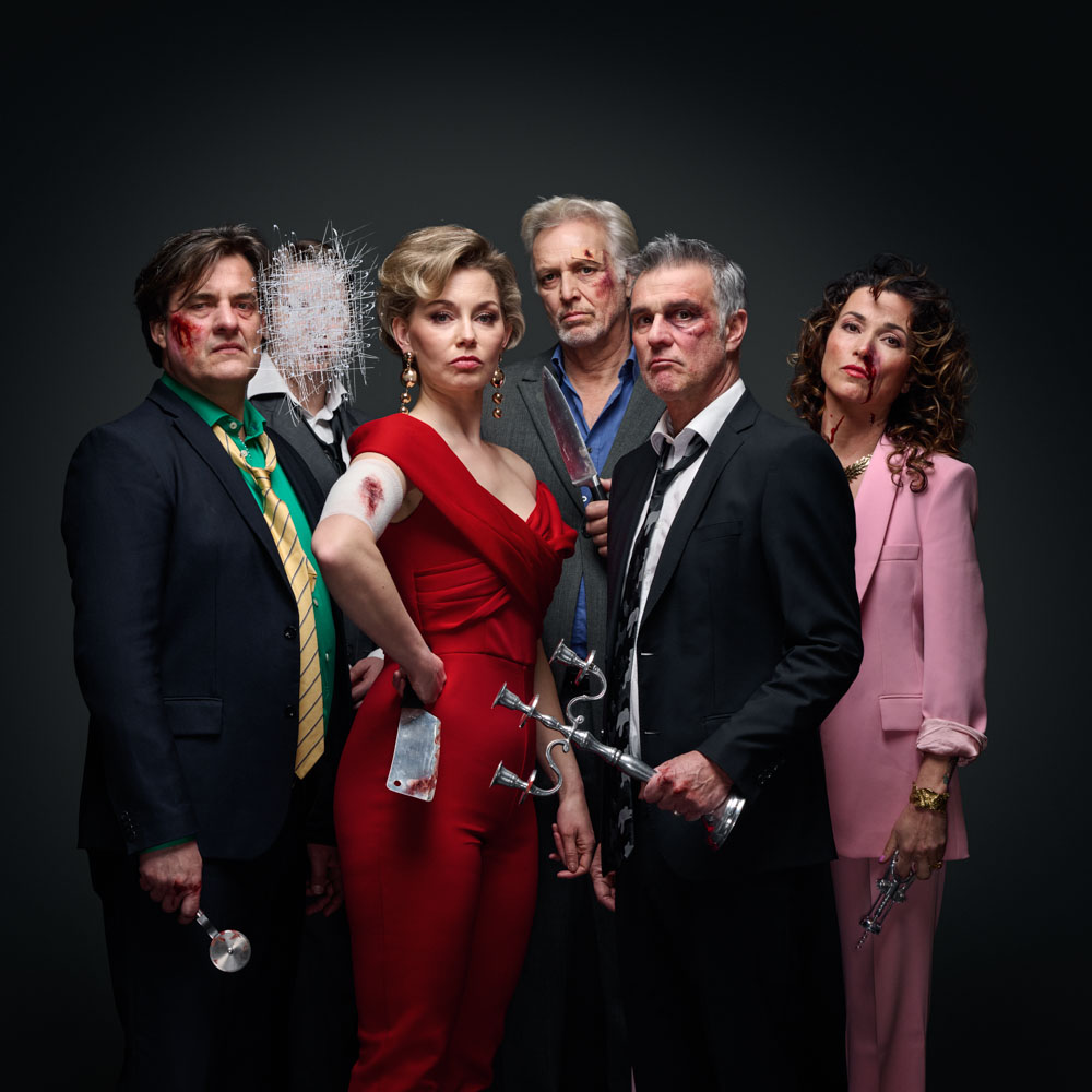 Groep spelers van De Nieuwe Ploeg met bloed op gezicht en kleding kijkt ernstig in de camera, messen en kaarsenstandaard in beeld.