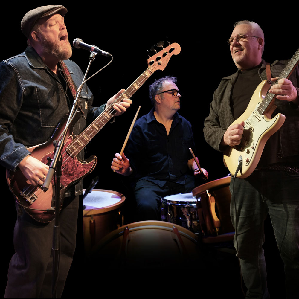 Drie muzikanten van Ocobar spelen gitaar, bas en drums op een donker podium.