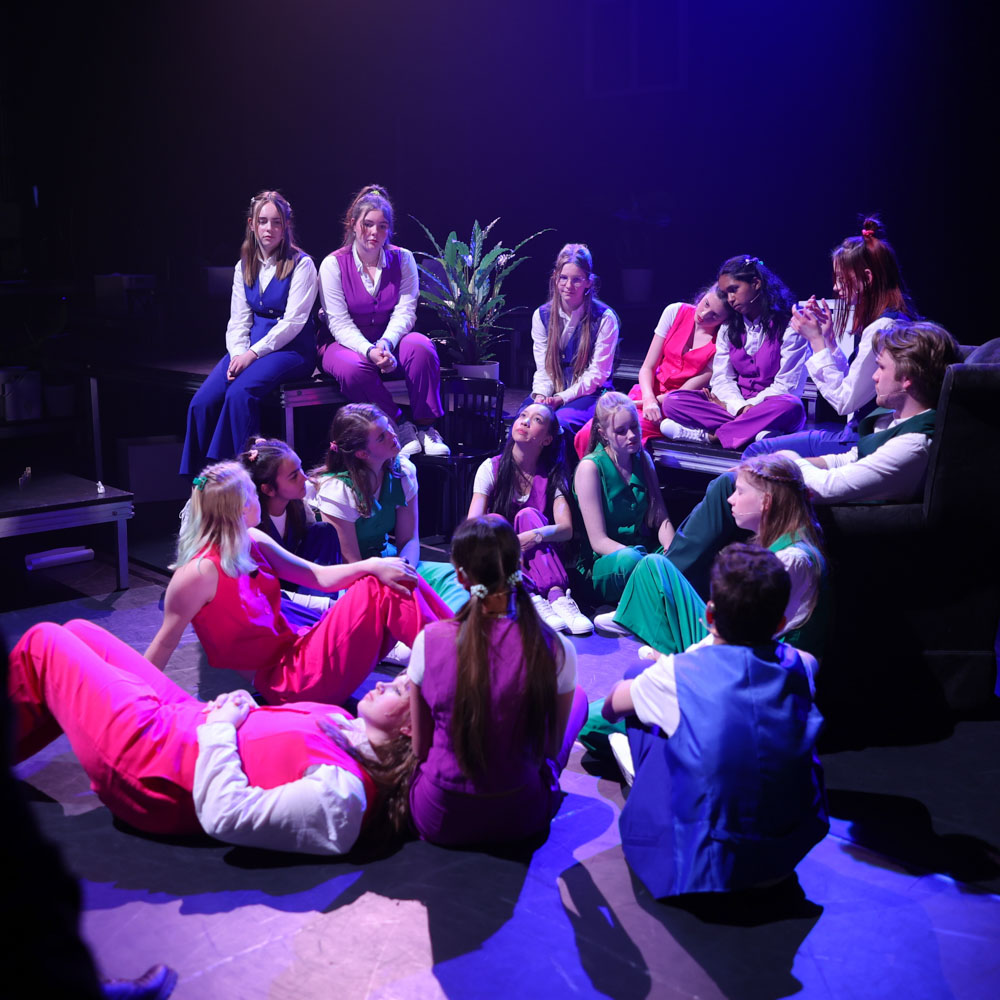 Groep jonge spelers zit in een kring op het podium onder paarsblauw theaterlicht.