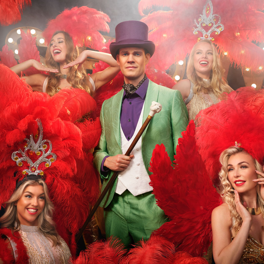 Tim Fransen in groen pak tussen showgirls met rode pluimen en glitterjurken.