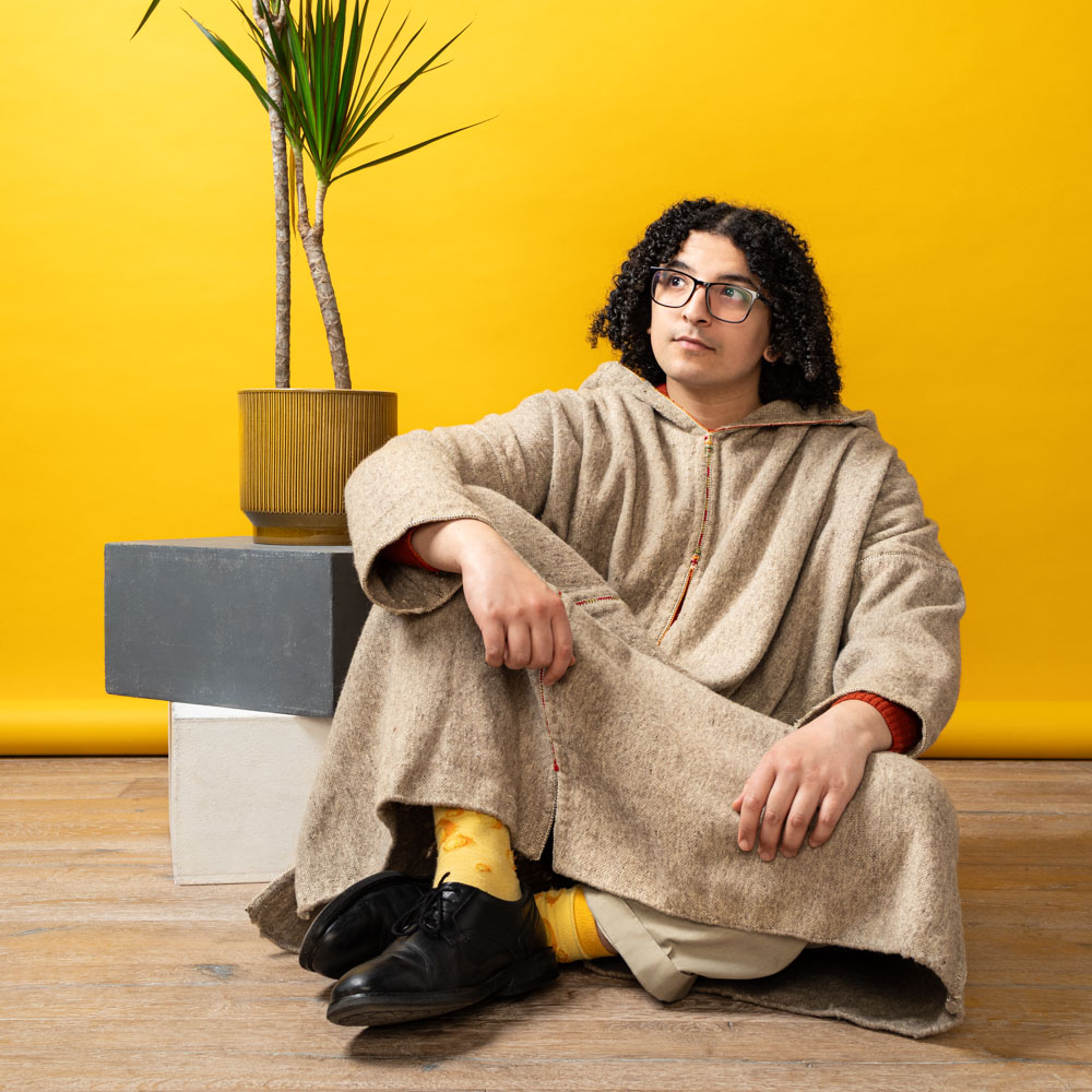 Ayoub zit in traditionele kleding op de grond voor een gele achtergrond met plant.