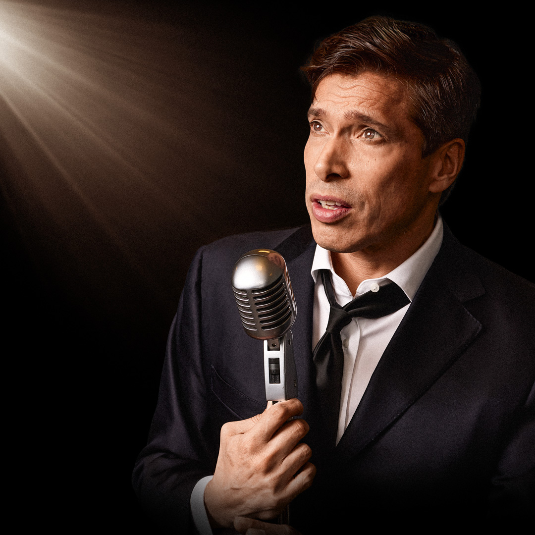 Zanger van BREL in pak houdt vintage microfoon vast onder warm spotlight.