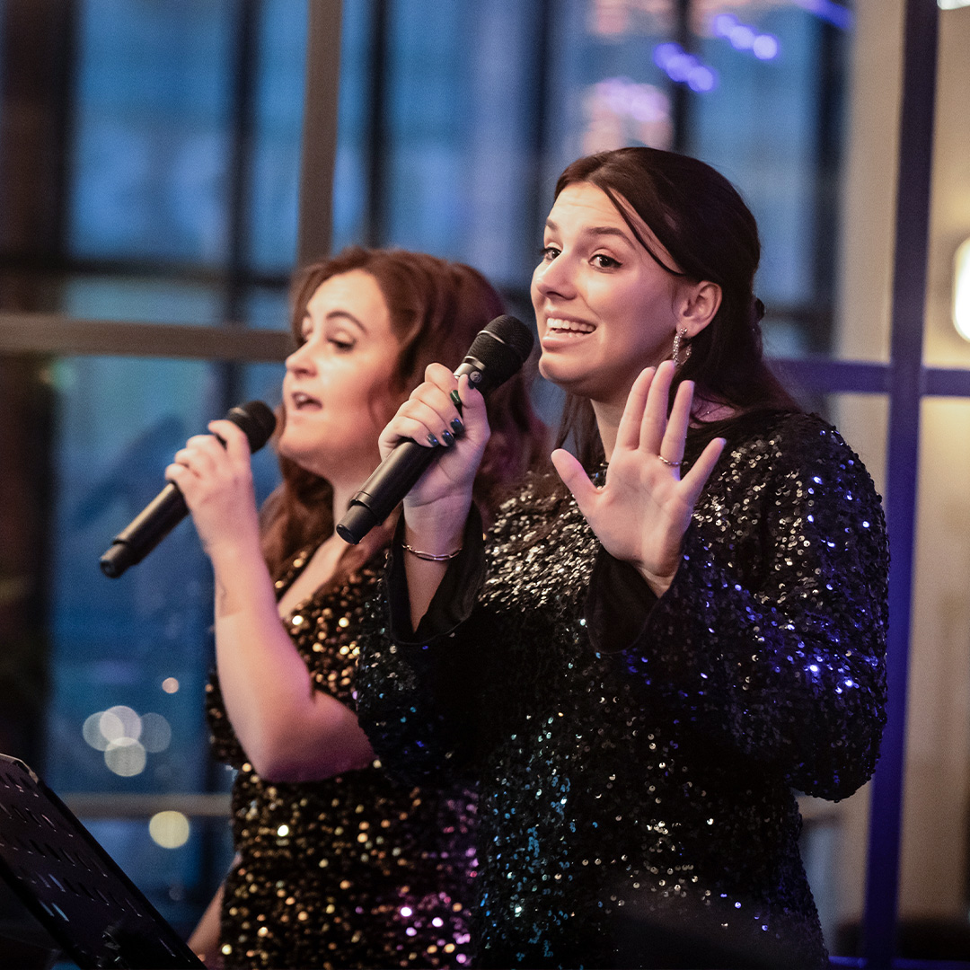 Twee zangeressen zingen enthousiast in glitterjurken tijdens De Muziekbingo.