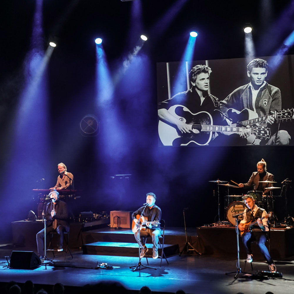 Muzikanten spelen onder blauw licht met oude foto van Everly Brothers op scherm.