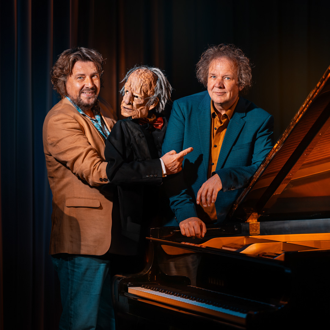 Fred Delfgaauw met pop en Jeroen van Veen bij vleugel voor donker gordijn.