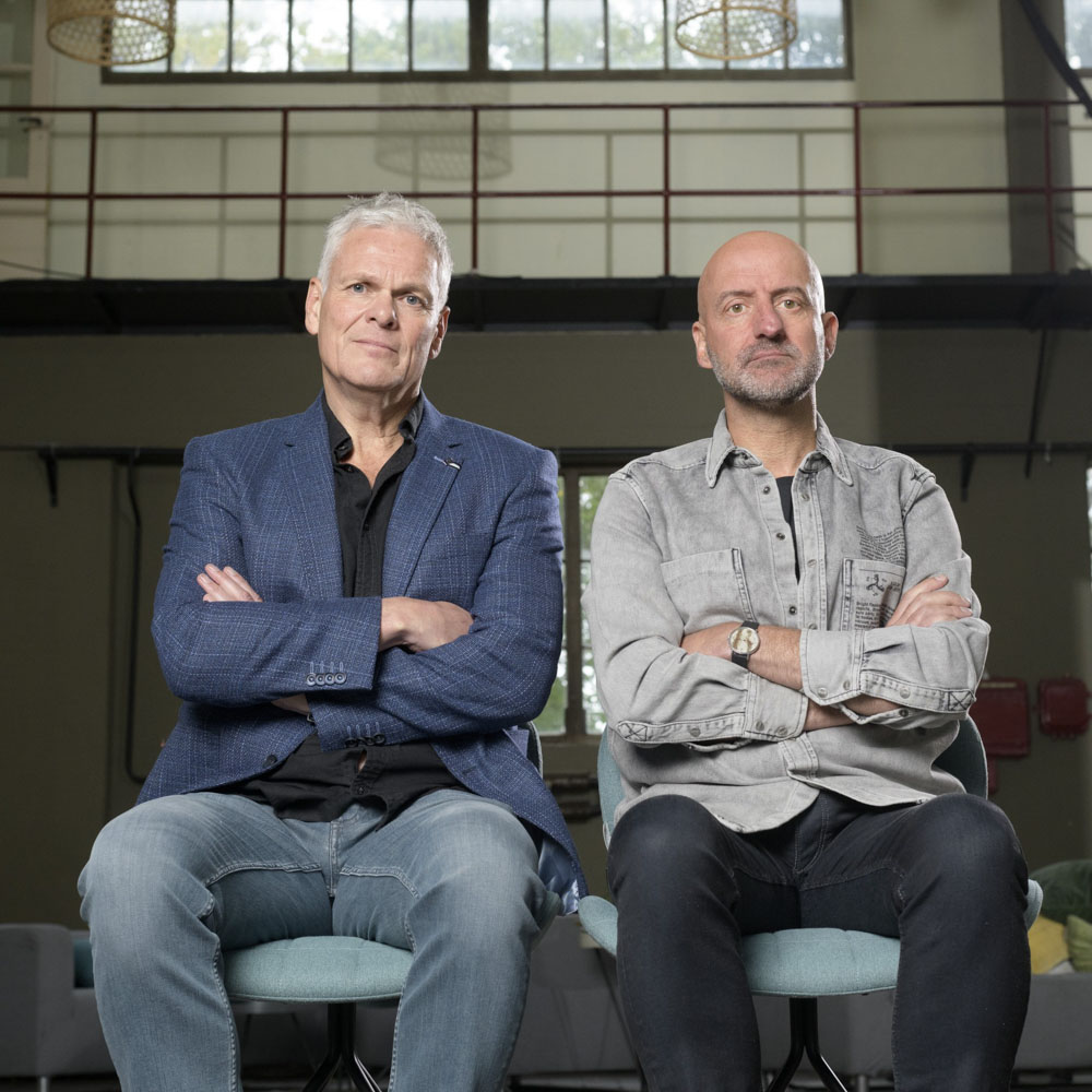 Joep en Rob zitten met armen over elkaar op stoelen in een industriële ruimte.