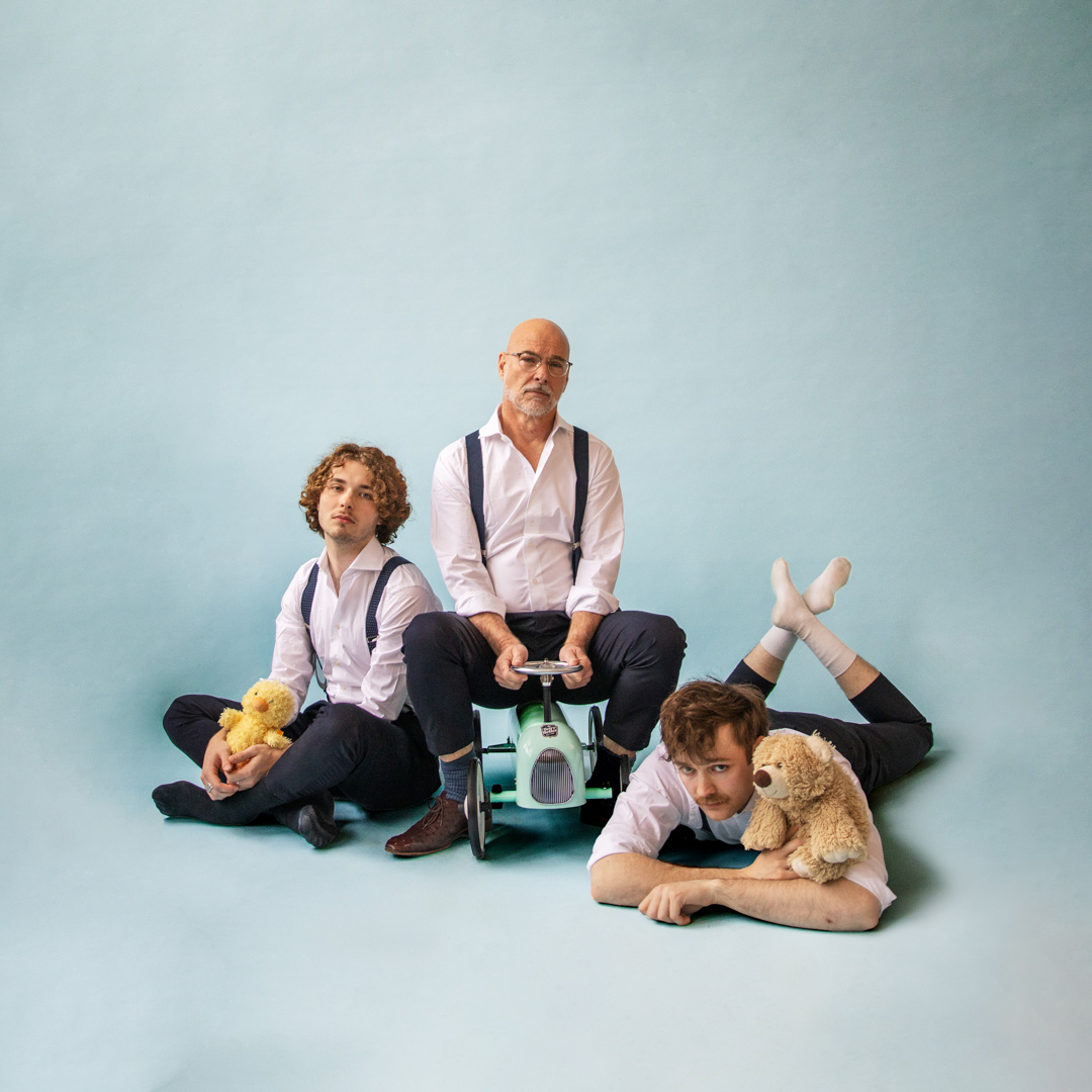 Rob Kamphues zit serieus op speelgoedauto tussen twee mannen met knuffels.