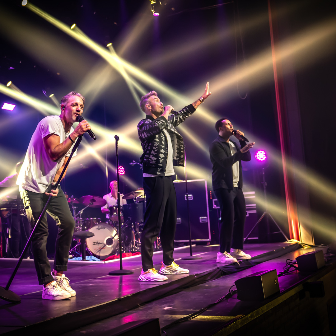 Boyband zingt op rij met kleurrijke lichtstralen en liveband op de achtergrond.