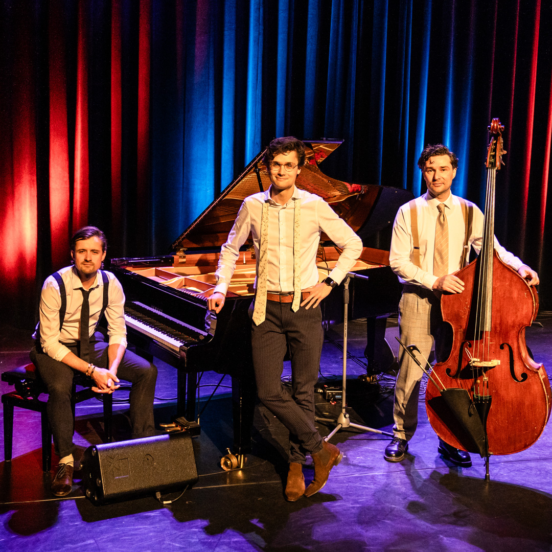 Dennis van Aarssen staat voor de vleugel tussen zijn pianist en contrabassist op een verlicht podium.