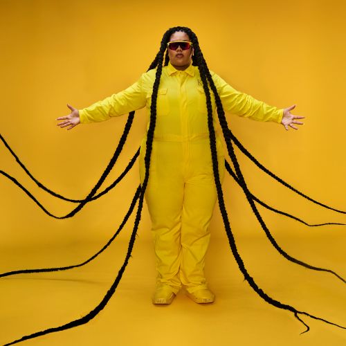 Isabelle Kafando spreid haar armen en handen uit in een gele outfit voor een gele achtergrond. Zij heeft een donkere bril op en meerdere vlechten die langs haar heen bewegen.